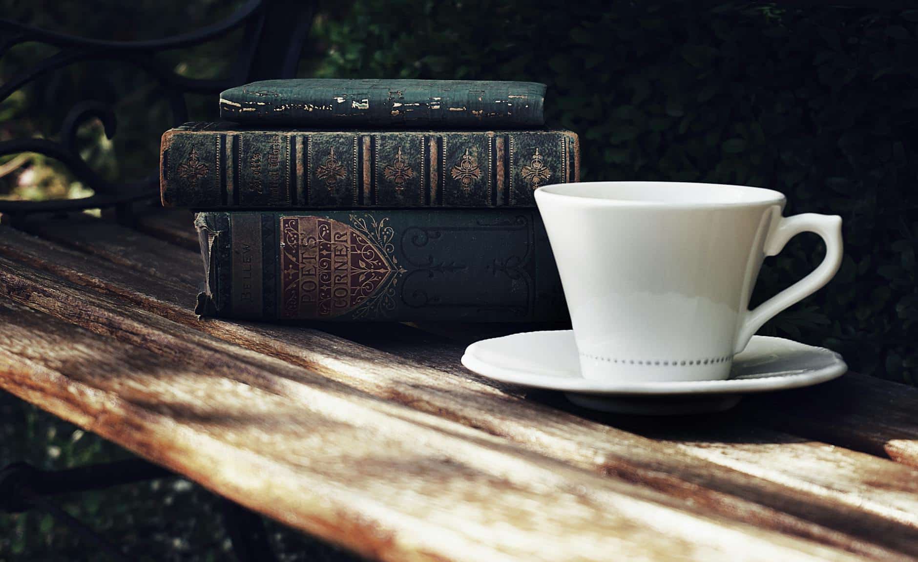 photo of ceramic cup and saucer next to a pile of books on wooden park bench